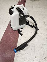 White plastic backpack sprayer with black straps and connected hose and spray gun on floor with white tile and brown tile pattern.