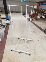 Front view of two white collapsible garment racks standing side by side on a tiled floor in a store with caster wheels visible at base.