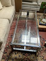 Rectangular coffee table with clear glass top and aluminum frame. Positioned next to a beige couch on patterned carpet.