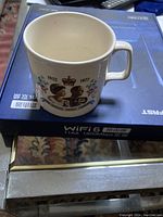 Photo of ceramic cup with Queen Elizabeth II and Prince Philip portraits, crown and floral decoration, next to a box