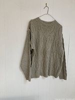 Beige knit cardigan hanging on a white wall, showing full front view with cable knit pattern.