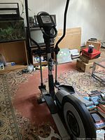 Front view of the black and gray elliptical exercise machine in a carpeted living room setting with surrounding boxes and furniture.