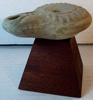 Front side view of light brown clay oil lamp on dark mahogany pyramid base highlighting spout and central hole.