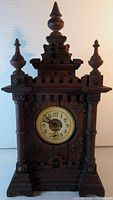 Front view of wooden steeple mantle clock with ornate carved details and clock face showing Arabic numerals and alarm dial.