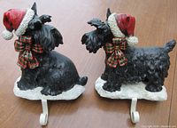 Pair of large black Scottie dog stocking holders wearing red Santa hats and plaid bows, viewed side-by-side on a wooden surface, showing the full figures, bases and white metal stocking hooks.