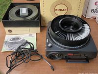 Kodak Carousel 800 slide projector loaded with a slide wheel and an additional slide wheel in box alongside instruction manuals and slides on table.