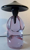 Front view of the pink glass figure with black hat and hair showing the sitting pose and clear pink glass body.
