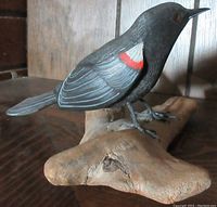Side angle of the hand carved and painted Red Winged Black Bird perched on a natural wood base, showing red wing markings and orange eyes.