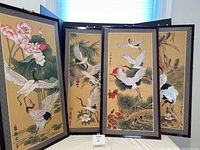 Four Asian silk screen painting panels displayed standing, showing cranes among floral and natural scenes with calligraphy, set against a beige background with dark borders.