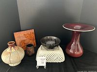 Collection of ceramic and pottery items: tall maroon pedestal vase, brown and beige drip glaze vases, black pottery bowl, white woven basket, and burnt orange ceramic trivet.