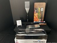 Photo showing the full cutlery set in plastic package with knives on display along with meat cleaver, grill spatula, grilling planks, and tool bag