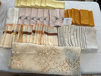 All cloth napkins and vintage Quaker Lace table linen laid out on a table. Different styles and colors arranged in groups: yellow, floral embroidered, mustard yellow, striped, beige linen, and folded lace table linen.