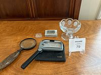 Displays a clear glass compote with scalloped rim, an antique magnifying glass with a wooden handle, a modern black plastic Bausch & Lomb magnifier with light, a coin in a round protective holder, and a rectangular marble block with a 10-cent metal stamp mounted on it.