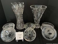 Five pieces of cut glass including three candy jars with lids and two decorative vases against a black background.
