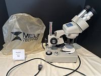 Full side view of Meiji EMT stereo microscope with attached power cord and plastic protective cover behind it.