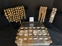 Photo showing multiple strings of large metallic beads with perforations displayed upright, and a compartmentalized box containing numerous small loose metal beads in various sizes and finishes.