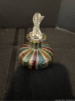 Photo of a rounded perfume bottle with multicolored stripes and a twisted clear glass stopper on top, displayed on a black textured surface.