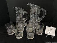 Two etched floral glass pitchers, one large with scalloped rim and one smaller, along with six matching etched floral drinking glasses arranged in front.