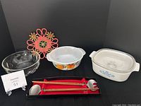 Photo of assortment of vintage serving bowls including glass, Fire King, and Corning pieces, red woven trivet, and boxed teak wood and stainless steel serving cutlery set.