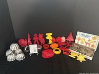 Wide view showing assortment of red, yellow plastic vintage cookie cutters and mini aluminum Jello molds, plus boxed holiday cookie cutter set