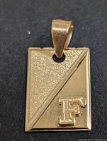 Top-down view of rectangular gold pendant displaying contrasting textured surfaces divided diagonally and embossed letter 'F'.