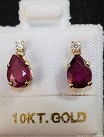 Front-facing view of pair of 10K gold earrings with pear-shaped ruby and small round moissanite stones on white holder stamped 10KT GOLD.