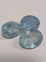 Three faceted aquamarine gemstones on plain white background, two oval cut and one pear cut, light to medium blue with inclusions visible.