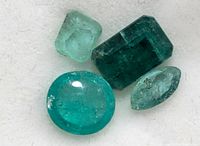 Close-up photo of four emerald gemstones in varied cuts and shades of green showing natural inclusions, taken on white background.