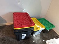Four large black heavy duty plastic storage bins stacked with two lids on top (red and yellow lids).