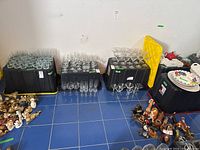 Five heavy duty black plastic storage bins with yellow lids arranged against wall; assorted glassware and decorative items on and around them for display only.