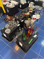 Overview of four large black heavy duty storage bins with assorted glassware and kitchen items on top and around them in a room with blue tiled floor.