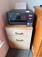 Printer on top of wooden cabinet showing front control panel, ink tanks, drawer handles and surrounding area.