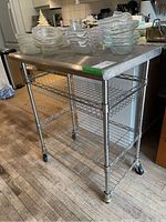 Full view of the metal baker style cart with stainless steel top and wire shelves on caster wheels in a kitchen environment.