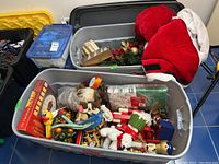 Full view of gray plastic bins filled with assorted Christmas decorations including stockings, figurines, and mantel decor.