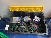 Black plastic bin with yellow lid containing tangled green wire Christmas lights and three NOMA boxes of LED decorative bulbs in colors red, orange, yellow, green, blue.