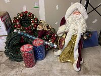 View of large Santa figurine with burgundy velvet coat trimmed in white fur, wreaths, ornaments containers and wrapping paper on concrete floor