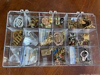 Clear plastic jewelry display case containing assorted small pins, badges, rings, some in plastic bags.