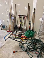 Full lot view showing various long handled garden hand tools including spades, rakes, hoe, brooms, two green watering cans, a Black and Decker electric hedge trimmer, pruners, hacksaw, garden hose.