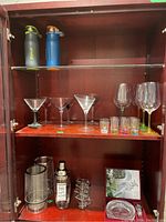 Overview of glassware and barware items displayed on wooden cabinet shelves including martini glasses, wine glasses, shot glasses, metal cocktail shaker, wine chillers, and glass holder stand.