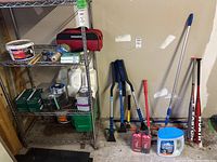 Wide angle of lot showing metal shelf with supplies and collection of cleaning brushes, squeegees, baseball bats, and containers on floor