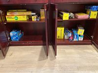 Wide view of kitchen cabinet opened showing various boxes of kitchen wraps, storage bags and trash bags on two shelves.