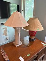 Two table lamps on wooden table, one with ceramic column-style base and beige fabric shade, the other with decorative ceramic and metal base with red and gold design and pleated fabric shade