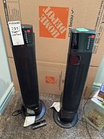 Pair of Lasko tower heaters standing on carpet in front of a Home Depot box with one remote on the floor