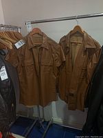 Two brown leather garments hanging side by side on a rack: short-sleeve shirt and long-sleeve jacket