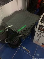 Stack of black rubber interlocking floor mats with puzzle-like edges and diamond plate texture, approximately 22x22 inches each.