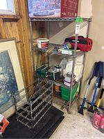 Metal utility shelving unit with four wire shelves showing some wear, placed against a wall in a garage setting with various items on and around it.