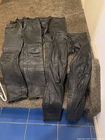 Three pairs of black leather pants laid flat showing front sides, indicating fabric texture, sizes, and details like pockets and belt loops.