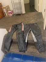 Two pairs of black leather pants laid side by side on carpeted stairs showing overall style and color accents