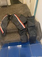 Two pairs of black leather motorcycle pants displayed on carpet with one showing red, white, and blue side stripes and quilted padding.