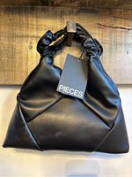 Front view of black Pieces purse with original tag, showing twisted handle and geometric front panel stitching.